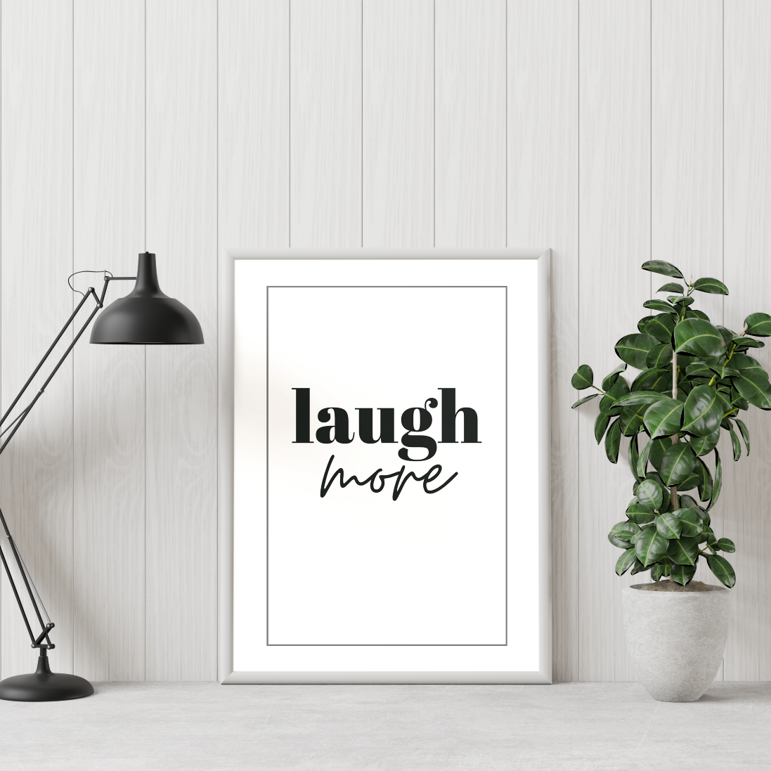 Poster Laugh more inramad mot vägg, till höger syns en grön växt och till vänster en skrivbordslampa