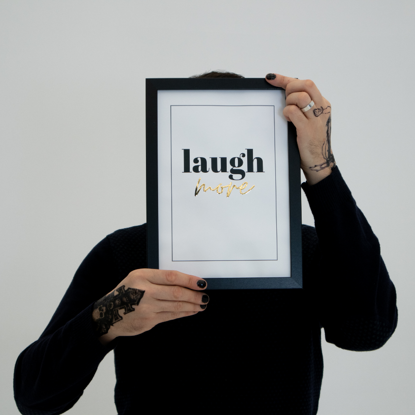 Poster Laugh More gold texten "more" i guldfoliering, inramad, en person med tatuerade händer håller upp tavlan framför sig så att ansiktet skyms.