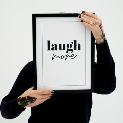 Poster Laugh more inramad hålls upp av en person med tatuerade händer, ansiktet skyms av postern