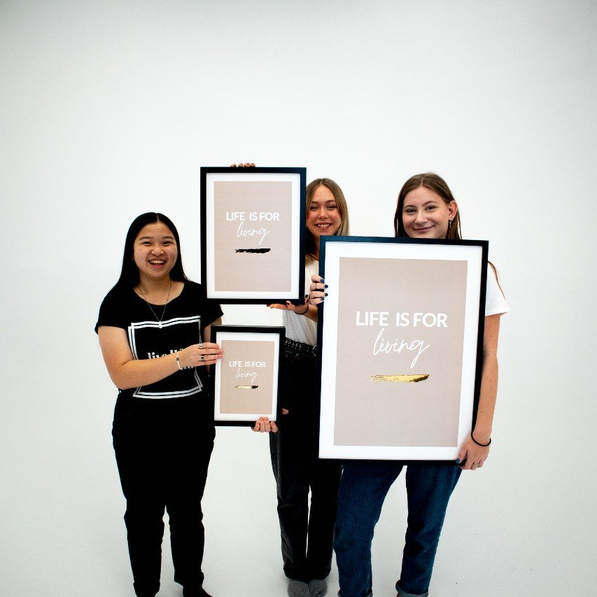 Poster Life is for living, med guldfoliering, alla tre storlekar hålls upp av tre unga kvinnor som ler