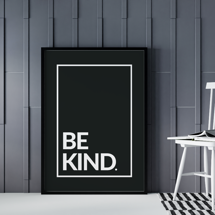 Poster Be kind, vita bokstäver mot svart bakgrund. Inramad och stående på golvet lutad mot grå vägg. En pinnstol syns till höger