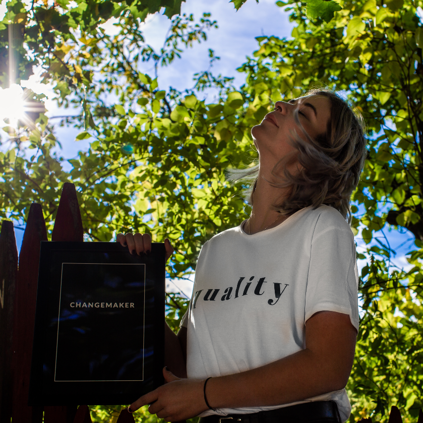 Poster Changemaker inramad. Hålls upp utomhus av en person som vänder ansiktet mot solen. Personen är klädd i vit T-shirt med texten equality
