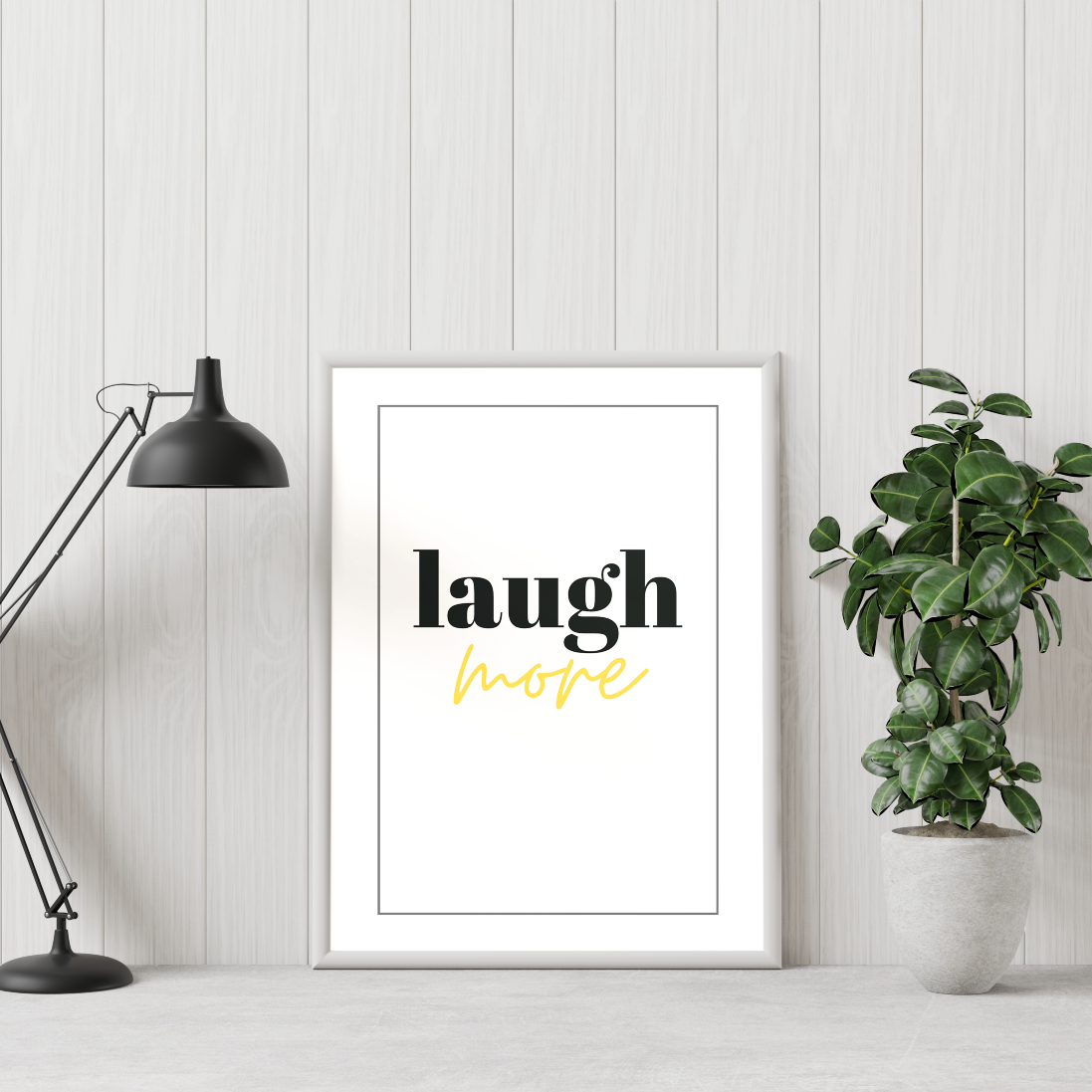 Poster Laugh More gold texten "more" i guldfoliering, inramad, till höger syns en grön växt och till vänster en skrivbordslampa