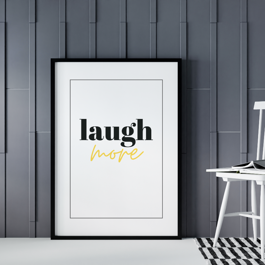 Poster Laugh More gold texten "more" i guldfoliering, inramad mot grå vägg, en vit stol syns til lhöger