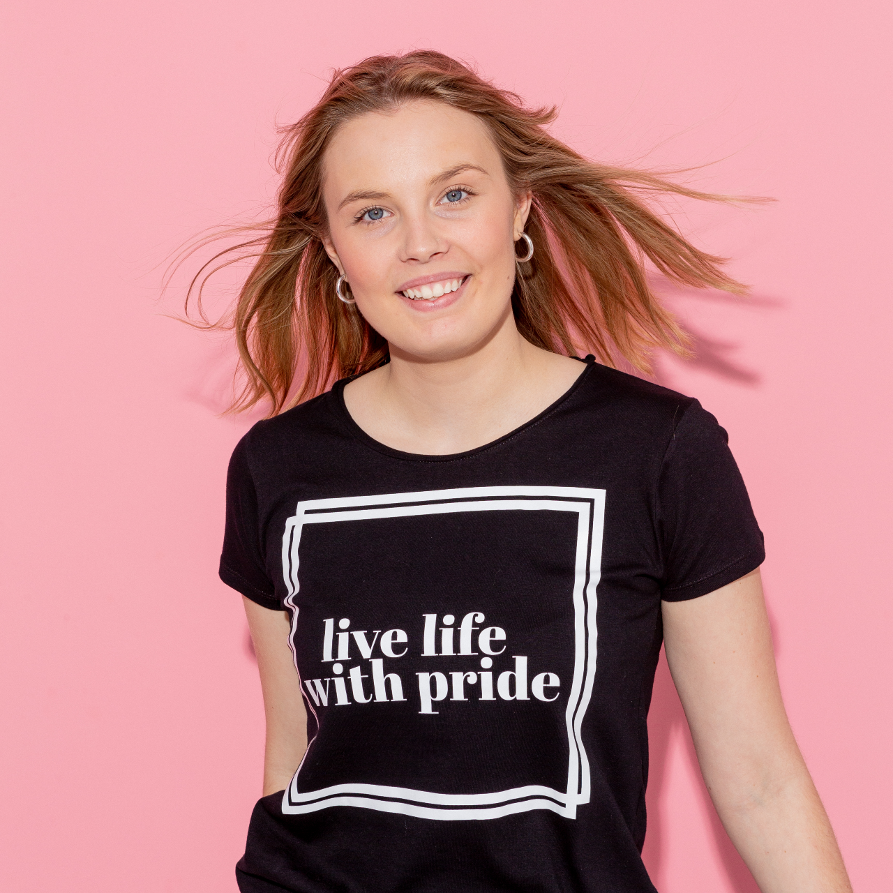 T-shirt Live Life, modell i rörelse framåt, håret blåser bakåt. Rosa bakgrund