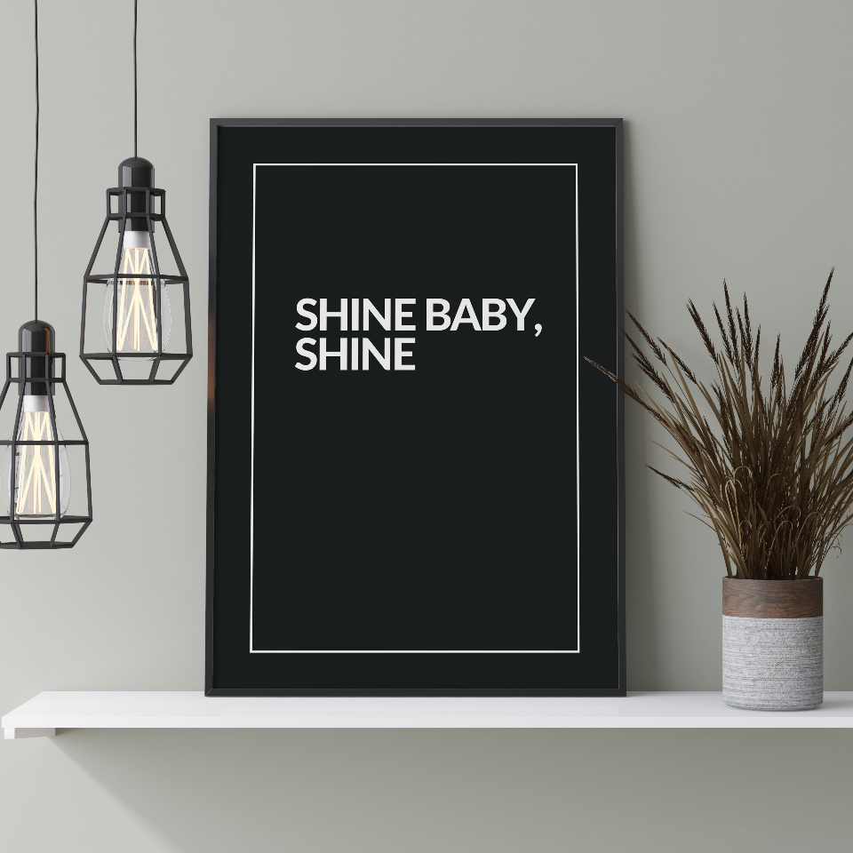Poster Shine, inramad stående på hylla med grön växt till höger och två lampor hängandes till vänster