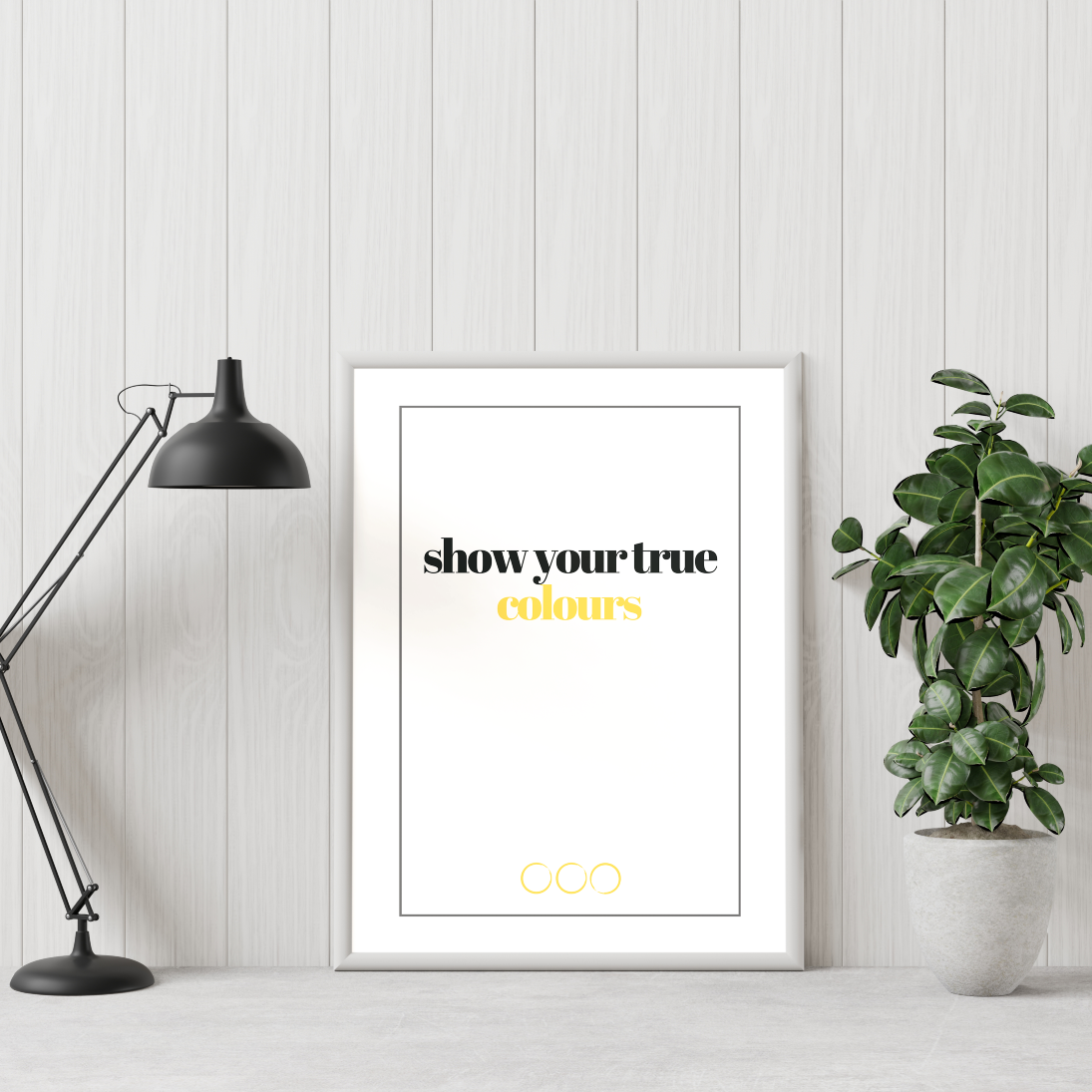 Poster True colours, gold. Texten "colours" i guldfoliering. inramad lutad mot  vit vägg. Grön växt till höger och skrivbordslampa till vänster