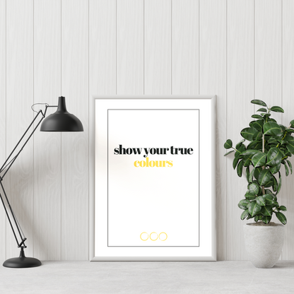 Poster True colours, gold. Texten "colours" i guldfoliering. inramad lutad mot  vit vägg. Grön växt till höger och skrivbordslampa till vänster